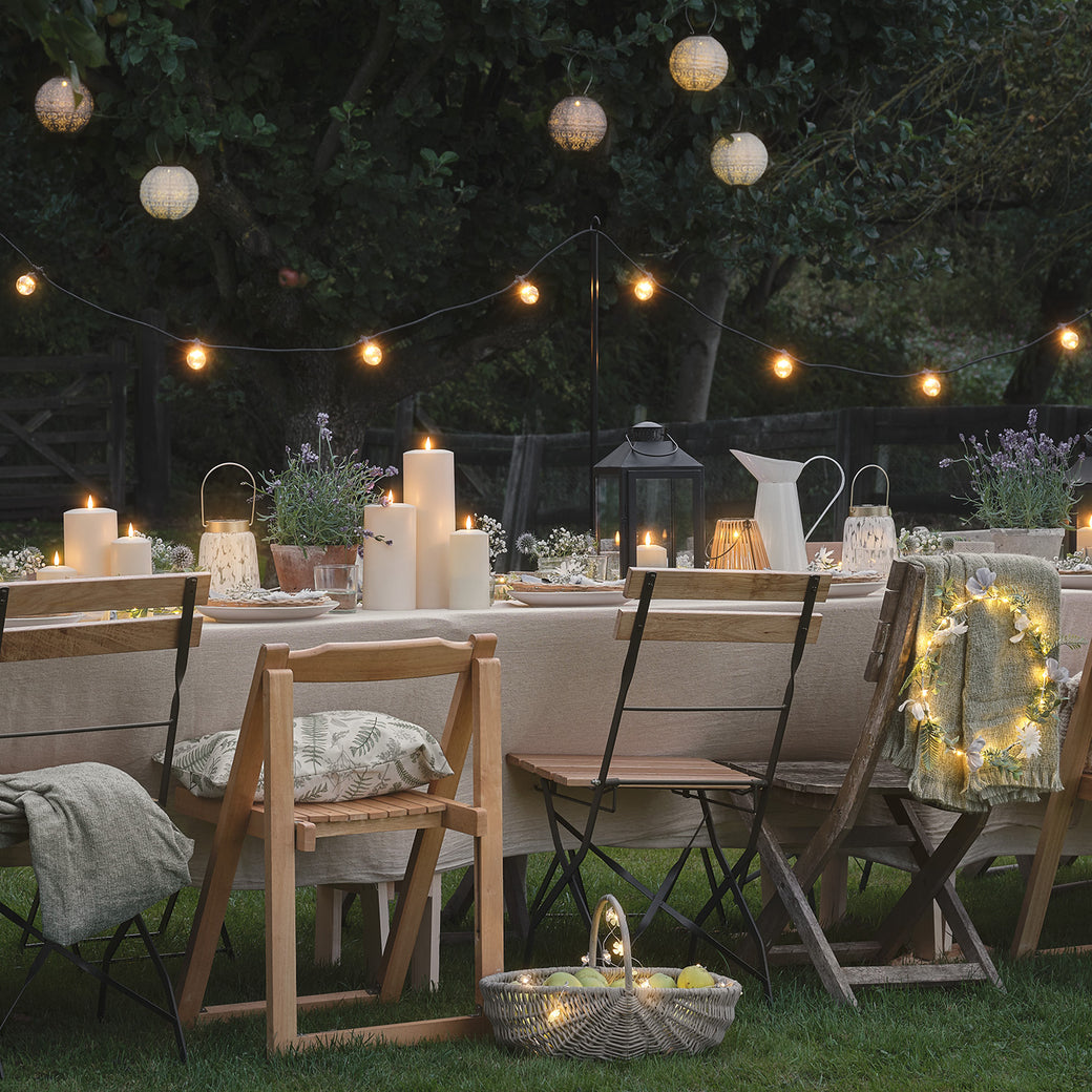 Outdoor dining table set with LED candles and lanterns, with festoon lights and fabric lanterns hanging overhead.