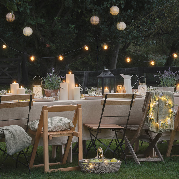 Outdoor dining table set with LED candles and lanterns, with festoon lights and fabric lanterns hanging overhead.