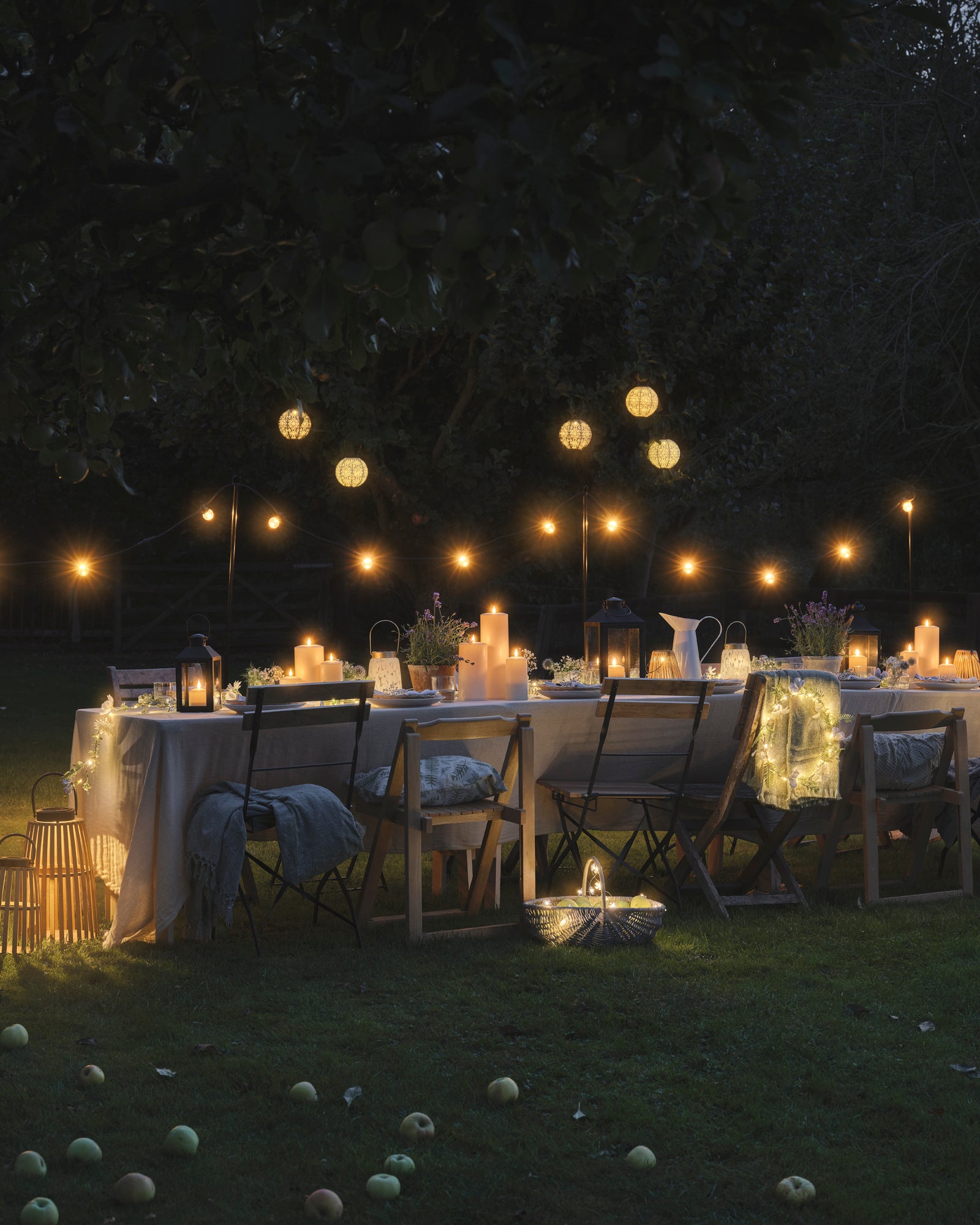 An outdoor dining table set with LED candles and lanterns, underneath hanging lanterns and festoon lights