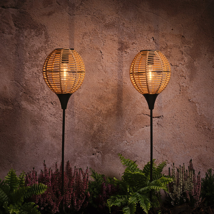 Two glowing wicker solar stake lights positioned in a flower bed against a wall