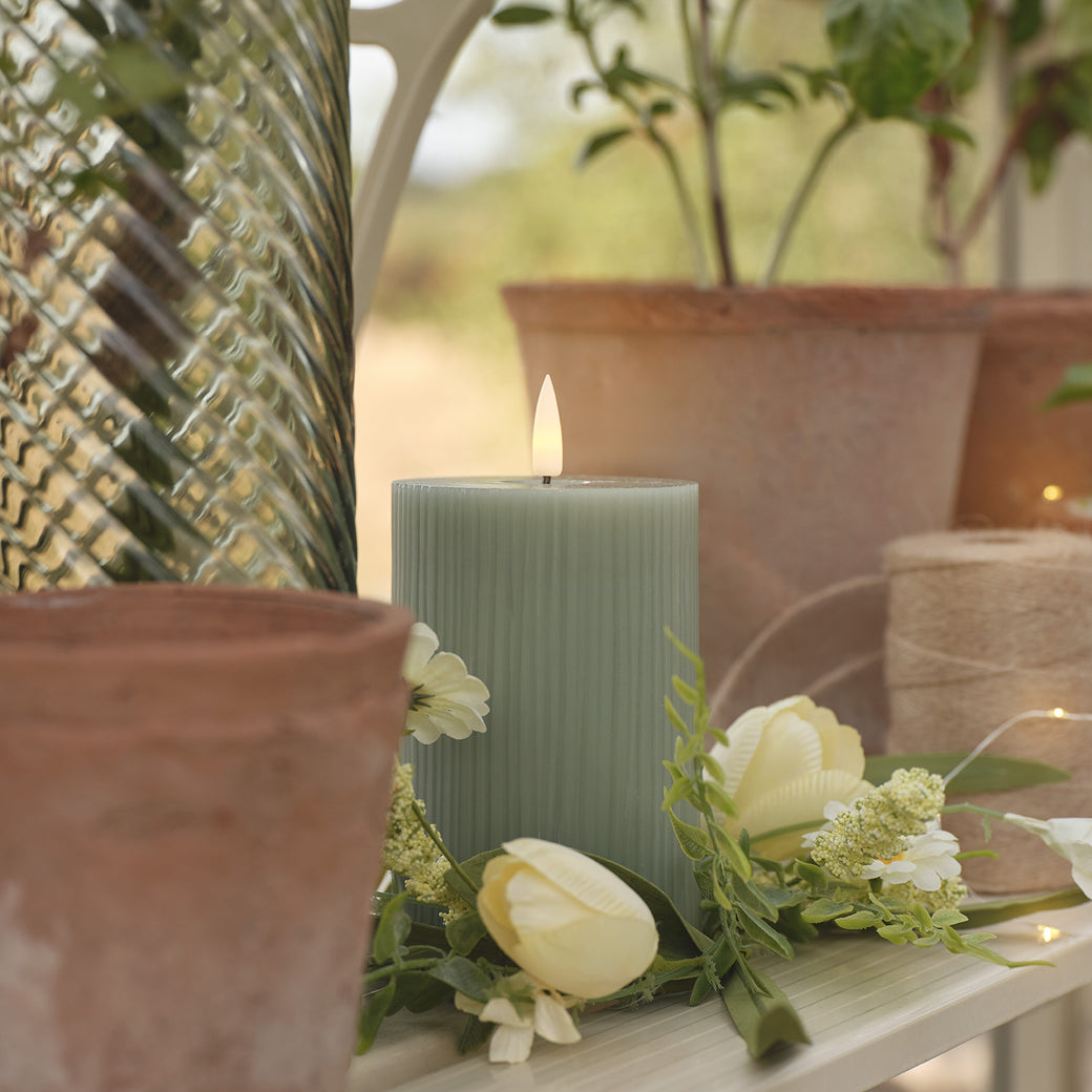 A sage green LED candle on a greenhouse shelf