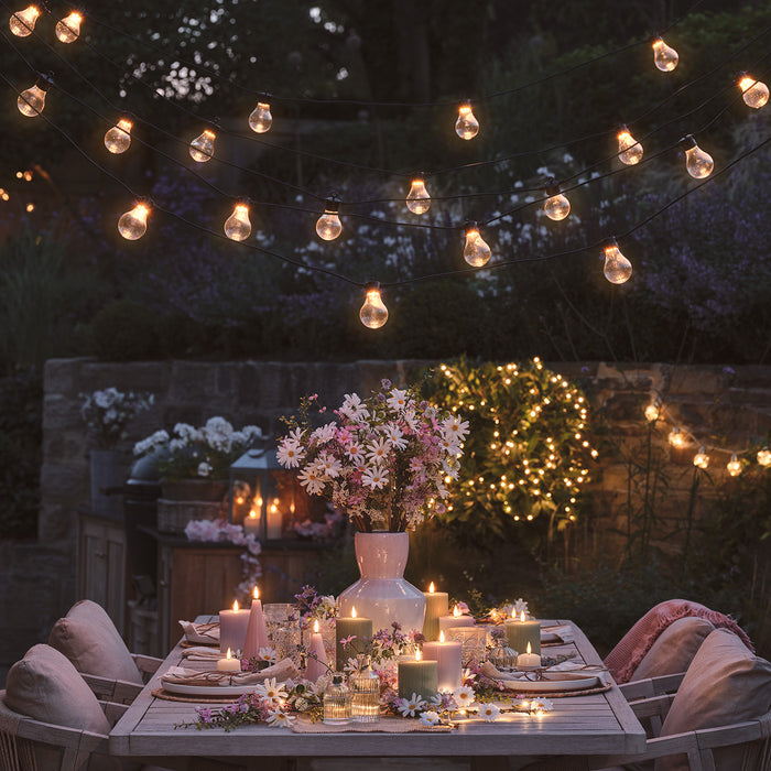 Outdoor dining table at night with candles and a bouquet of pink and white flowers on it with festoon lights hanging above it
